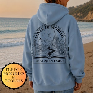 Può includere: Felpa con cappuccio in pile azzurro con una grafica sul retro che recita "LOVER OF WORLDS" sopra un'illustrazione di un castello, montagne e un libro aperto. Il testo "THAT AREN'T MINE" è sotto l'illustrazione. La felpa è mostrata su una persona sulla spiaggia.