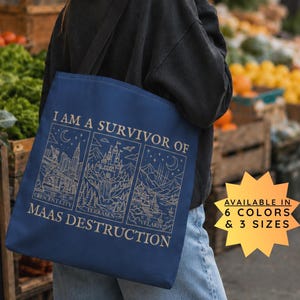 Pode incluir: Uma bolsa tote azul-escura com um gráfico de linhas claras que mostra três painéis com paisagens urbanas e naturais. O texto diz I AM A SURVIVOR OF MAAS DESTRUCTION, com as etiquetas Crescent City, Terrasen e Velaris sob cada painel.