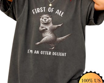 May include: Dark gray t-shirt with the text "FIRST OF ALL I'M AN OTTER DELIGHT" and a graphic of an otter. The shirt is made of 100% US cotton and has a Comfort Colors label.