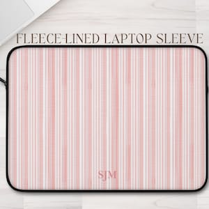 May include: A fleece-lined laptop sleeve with a pink and white striped pattern. The sleeve has a black border and a zipper closure. The initials "SJM" are visible on the lower right corner. The text "FLEECE-LINED LAPTOP SLEEVE" is printed at the top.