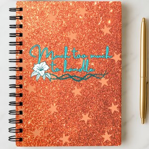 May include: A notebook with a glittery orange cover, peach stars, and the teal text "Much too much to handle". A gold pen lies next to the notebook. The notebook has a black spiral binding.