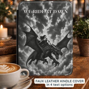 May include: A black and white faux leather Kindle cover featuring a graphic illustration of a person riding a dragon against a cloudy sky. The text WE RIDE AT DAWN is printed in white at the top. The cover has a textured surface and rounded corners.