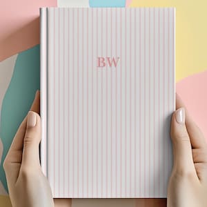 Op de afbeelding: Een hardcover notitieboek met een roze en wit gestreept patroon. De initialen "BW" staan in zachtroze op de kaft. Het notitieboek wordt door twee handen vastgehouden tegen een kleurrijke, abstracte achtergrond.