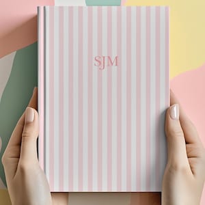 May include: A hardback notebook featuring a vertical striped pattern in alternating shades of light pink and white. The centre of the front cover displays the embossed capital letters SJM in a simple, clean serif font, centred vertically and horizontally.