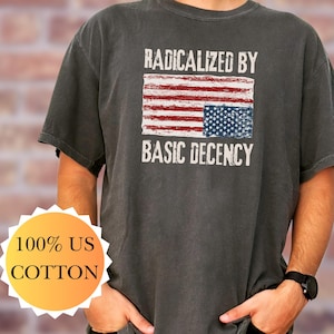 Op de afbeelding: Donkergrijs t-shirt met de tekst "RADICALIZED BY BASIC DECENCY" boven een ontwerp van de Amerikaanse vlag. Het shirt is gemaakt van 100% US katoen.