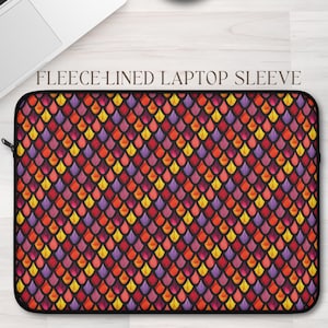 May include: A fleece-lined laptop sleeve with a colorful scale pattern in shades of red, orange, yellow, and purple. The text "FLEECE-LINED LAPTOP SLEEVE" is printed at the top. A laptop and a cup of coffee are in the background.