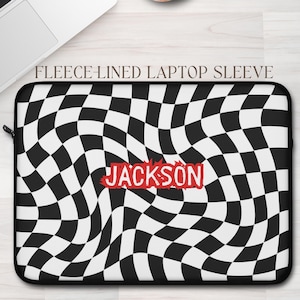 May include: Black and white checkered laptop sleeve with the name "JACKSON" in red. The sleeve is fleece-lined and has a zipper closure. A laptop and a cup of coffee are in the background.
