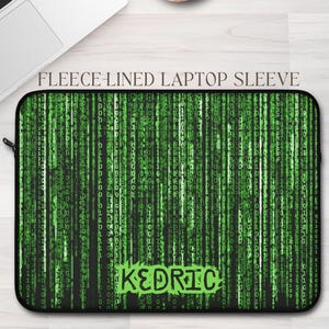 May include: Black laptop sleeve featuring a green digital rain design and the name "Kedric" in green. The sleeve is fleece-lined and has a zip closure. A laptop and a cup of coffee are also in the image.
