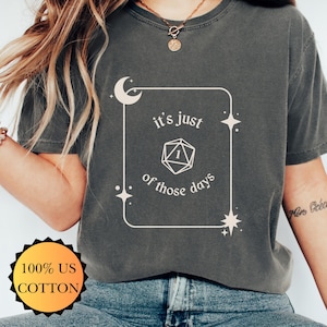 May include: Dark grey t-shirt with the text "it's just of those days" inside a white frame with a moon, stars, and a die graphic. The shirt is made of 100% US cotton.