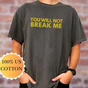 May include: Dark grey t-shirt with the yellow text "YOU WILL NOT BREAK ME". The shirt is made of 100% US cotton. The t-shirt has short sleeves and a crew neck.