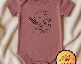 Puede incluir: Body de bebé rosa polvoriento con un dragón de dibujos animados negro sosteniendo una espada. La palabra "Huzzah" está impresa debajo del dragón. Una pegatina amarilla y negra dice "SOFT ROMPERS en 7 colores!"