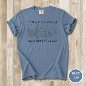 K&ouml;nnte beinhalten: Blaues Jeans-T-Shirt mit hochgekrempelten &Auml;rmeln. Der Aufdruck "I AM A SURVIVOR OF MAAS DESTRUCTION" befindet sich &uuml;ber drei Illustrationen von Stadt- und Landschaftsbildern. Das Shirt hat einen Rundhalsausschnitt und ein Etikett am Ausschnitt.