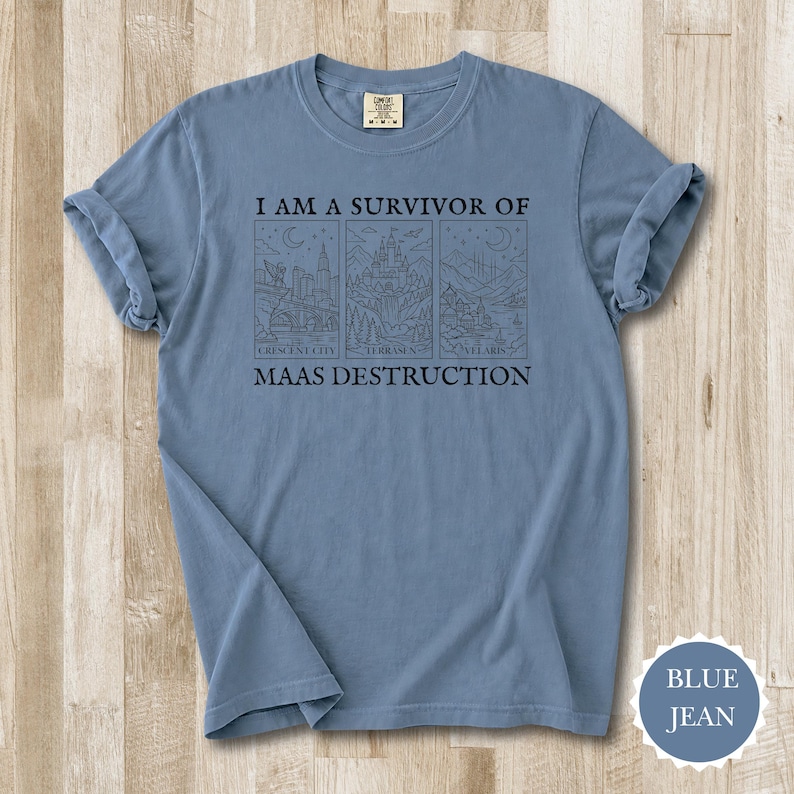 K&ouml;nnte beinhalten: Blaues Jeans-T-Shirt mit hochgekrempelten &Auml;rmeln. Der Aufdruck "I AM A SURVIVOR OF MAAS DESTRUCTION" befindet sich &uuml;ber drei Illustrationen von Stadt- und Landschaftsbildern. Das Shirt hat einen Rundhalsausschnitt und ein Etikett am Ausschnitt.
