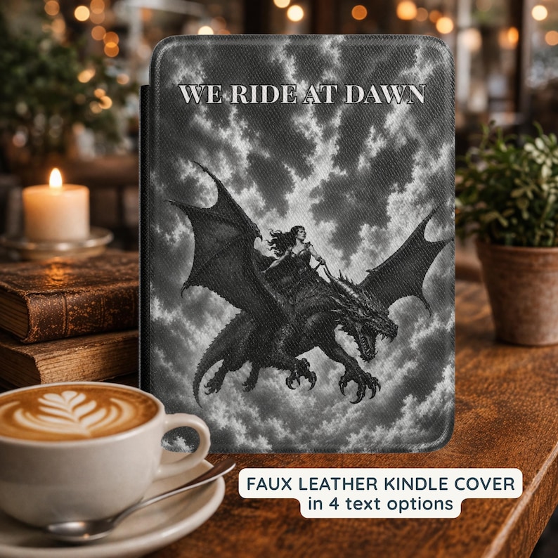 May include: A black and white faux leather Kindle cover featuring a graphic illustration of a person riding a dragon against a cloudy sky. The text WE RIDE AT DAWN is printed in white at the top. The cover has a textured surface and rounded corners.
