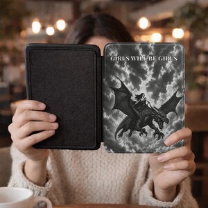 May include: A black e-reader case featuring a monochrome graphic of a figure riding a dragon against a cloudy sky. The text "GIRLS WILL BE GIRLS" is printed in white capital letters at the top. The case has a solid black interior lining.