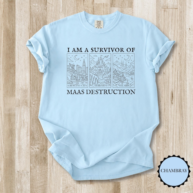K&ouml;nnte beinhalten: Hellblaues T-Shirt mit dem Text "I AM A SURVIVOR OF MAAS DESTRUCTION". Das Design zeigt drei illustrierte Tafeln mit Stadt- und Landschaftsmotiven. Das T-Shirt ist aus weichem Material.