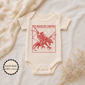 Puede incluir: Un body de beb&eacute; de manga corta en color crema con un estampado gr&aacute;fico rojo de una persona montando un drag&oacute;n. El texto WE RIDE AT DAWN aparece sobre la ilustraci&oacute;n. Una insignia circular a la izquierda indica NATURAL 100% cotton.