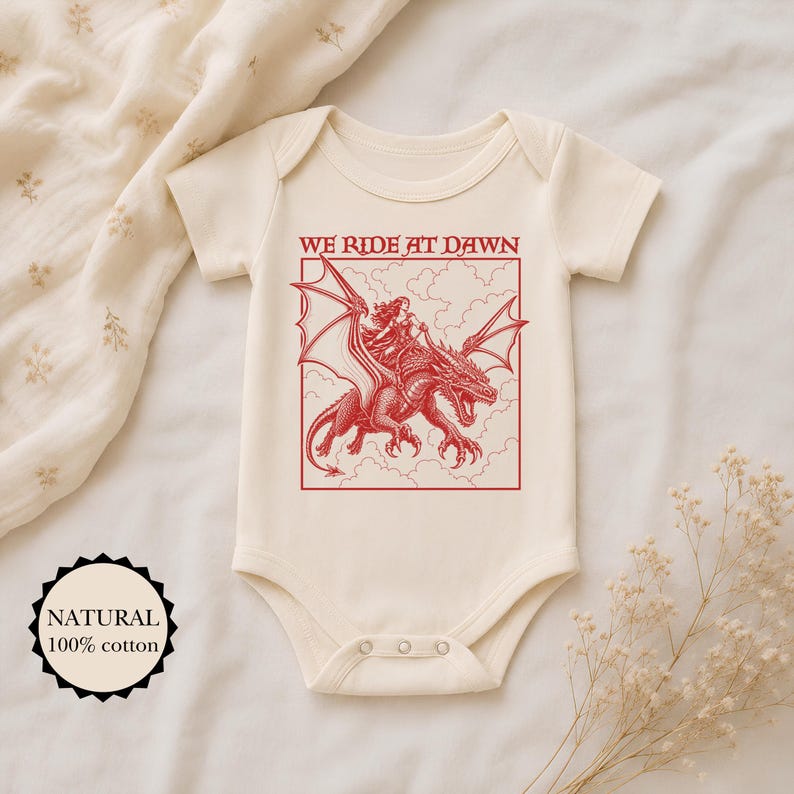 Puede incluir: Un body de beb&eacute; de manga corta en color crema con un estampado gr&aacute;fico rojo de una persona montando un drag&oacute;n. El texto WE RIDE AT DAWN aparece sobre la ilustraci&oacute;n. Una insignia circular a la izquierda indica NATURAL 100% cotton.