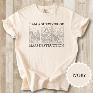 K&ouml;nnte beinhalten: Elfenbeinfarbenes T-Shirt mit dem Text "I AM A SURVIVOR OF MAAS DESTRUCTION" und drei illustrierten Tafeln. Die Tafeln zeigen Stadt- und Landschaftsbilder. Das T-Shirt ist gefaltet, mit sichtbarem &Auml;rmel und einem "IVORY" Farbetikett.