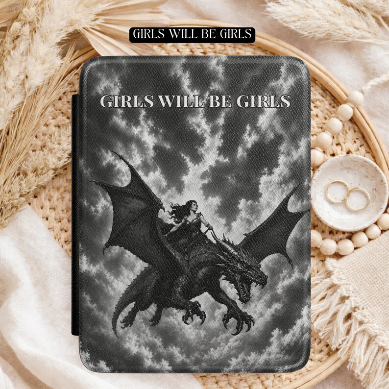 May include: A rectangular e-reader case featuring a black and white graphic of a person riding a dragon against a cloudy sky. The text "GIRLS WILL BE GIRLS" is printed in white capital letters at the top centre of the textured cover surface.