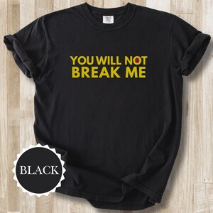 May include: Black t-shirt with the text "YOU WILL NOT BREAK ME" in yellow. A small red dot is above the letter "O" in "NOT". A black circle with the word "BLACK" in white is at the bottom left of the shirt.