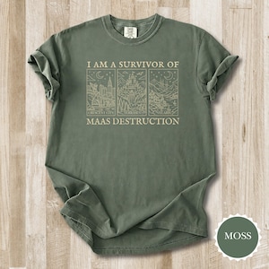 K&ouml;nnte beinhalten: Moosgr&uuml;nes T-Shirt mit hochgekrempelten &Auml;rmeln. Das Shirt zeigt den Text "I AM A SURVIVOR OF MAAS DESTRUCTION" &uuml;ber drei illustrierten Tafeln mit Stadt- und Landschaftsansichten. Das Shirt ist aus einem weichen, bequemen Material gefertigt.