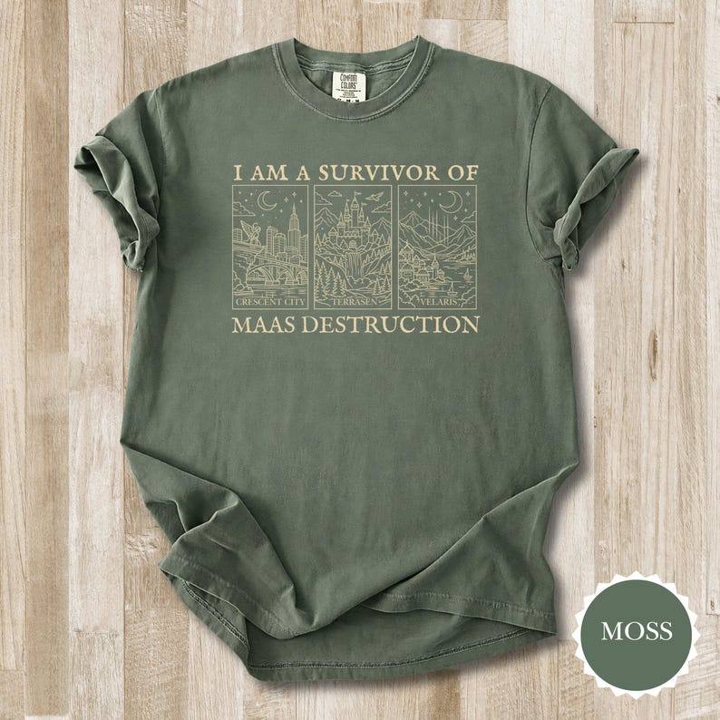 K&ouml;nnte beinhalten: Moosgr&uuml;nes T-Shirt mit hochgekrempelten &Auml;rmeln. Das Shirt zeigt den Text "I AM A SURVIVOR OF MAAS DESTRUCTION" &uuml;ber drei illustrierten Tafeln mit Stadt- und Landschaftsansichten. Das Shirt ist aus einem weichen, bequemen Material gefertigt.