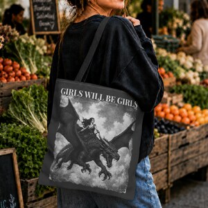 May include: A black tote bag featuring a monochromatic graphic print of a person riding a dragon with large wings. The text "GIRLS WILL BE GIRLS" is printed in white capital letters above the illustration, set against a cloudy, textured background.