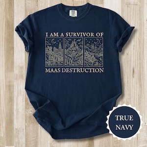 K&ouml;nnte beinhalten: Marineblaues T-Shirt mit dem Text "I AM A SURVIVOR OF MAAS DESTRUCTION" und drei illustrierten Tafeln. Die Tafeln zeigen Stadtlandschaften und Landschaften. Das T-Shirt hat einen Rundhalsausschnitt und kurze &Auml;rmel.