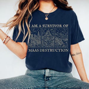 K&ouml;nnte beinhalten: Marineblaues T-Shirt mit wei&szlig;em Grafikdesign. Das Design zeigt drei Tafeln mit Illustrationen von Stadtlandschaften und Landschaften sowie den Text "I AM A SURVIVOR OF MAAS DESTRUCTION". Das Shirt ist ein bequemer, l&auml;ssiger Stil.