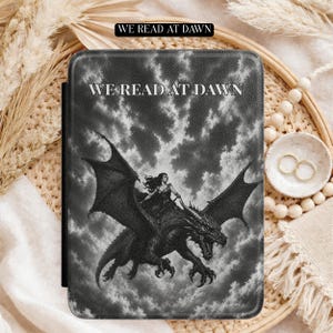 May include: A rectangular e-reader case featuring a black and white graphic of a person riding a dragon against a cloudy sky. The text WE READ AT DAWN is printed in white capital letters at the top centre of the cover, with a matching black label above.