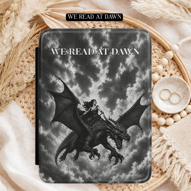 May include: A rectangular e-reader case featuring a black and white graphic of a person riding a dragon against a cloudy sky. The text WE READ AT DAWN is printed in white capital letters at the top centre of the cover, with a matching black label above.