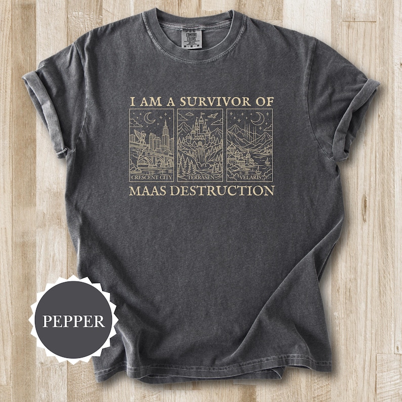 K&ouml;nnte beinhalten: Dunkelgraues T-Shirt mit dem Text "I AM A SURVIVOR OF MAAS DESTRUCTION" &uuml;ber drei Strichzeichnungen. Die Zeichnungen zeigen eine Stadt, ein Schloss und eine Berglandschaft. Die &Auml;rmel sind hochgekrempelt.