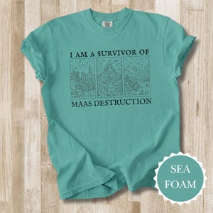 K&ouml;nnte beinhalten: T&uuml;rkisgr&uuml;nes T-Shirt mit dem Text "I AM A SURVIVOR OF MAAS DESTRUCTION" und drei illustrierten Tafeln. Die Tafeln zeigen Crescent City, Terrasen und Velaris. Das Shirt ist ein bequemer, l&auml;ssiger Stil.