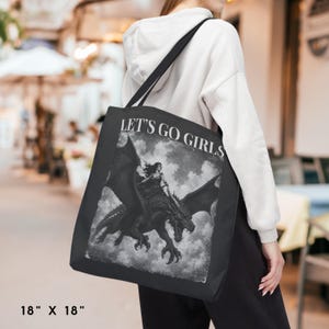 May include: A black tote bag measuring 18 inches by 18 inches featuring a white graphic print of a person riding a dragon. The text LET'S GO GIRLS is printed in white capital letters above the illustration of the dragon and rider in the clouds.