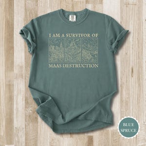 K&ouml;nnte beinhalten: Blau-Fichte-T-Shirt mit dem Text "I AM A SURVIVOR OF MAAS DESTRUCTION". Das Design zeigt drei Strichzeichnungen von Stadtlandschaften und Landschaften. Das Shirt liegt flach auf einer Holzoberfl&auml;che.