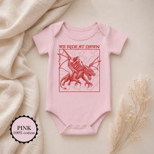 Puede incluir: Un body para beb&eacute; de color rosa claro con un estampado gr&aacute;fico rojo de una persona montando un drag&oacute;n dentro de un marco cuadrado. Sobre la ilustraci&oacute;n, el texto dice WE RIDE AT DAWN. La prenda tiene mangas cortas y cuello redondo.