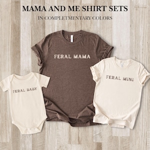 May include: A set of three matching shirts in complementary colors. The set includes a brown t-shirt with "FERAL MAMA" printed on it, a cream-colored t-shirt with "FERAL MINI", and a cream-colored baby onesie with "FERAL BABY". The text "MAMA AND ME SHIRT SETS" is at the top.