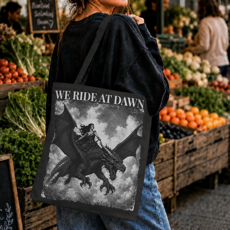 May include: A black tote bag featuring a white graphic print of a person riding a dragon with large wings. The text WE RIDE AT DAWN is printed in white capital letters above the illustration. The design has a distressed, textured appearance.