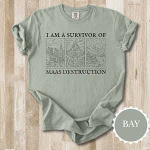 K&ouml;nnte beinhalten: Salbeigr&uuml;nes T-Shirt mit dem Text "I AM A SURVIVOR OF MAAS DESTRUCTION" &uuml;ber drei schwarzen Strichzeichnungen von Stadt- und Landschaftsbildern. Das Shirt hat hochgekrempelte &Auml;rmel und eine lockere Passform.