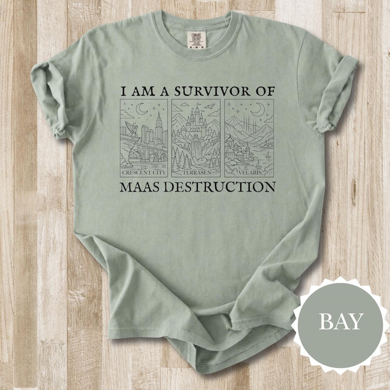 K&ouml;nnte beinhalten: Salbeigr&uuml;nes T-Shirt mit dem Text "I AM A SURVIVOR OF MAAS DESTRUCTION" &uuml;ber drei schwarzen Strichzeichnungen von Stadt- und Landschaftsbildern. Das Shirt hat hochgekrempelte &Auml;rmel und eine lockere Passform.