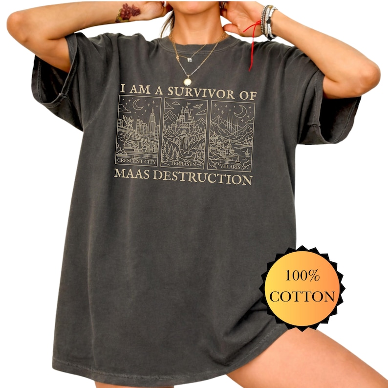 May include: Dark gray t-shirt with the text "I AM A SURVIVOR OF MAAS DESTRUCTION" and three illustrated panels. The panels depict cityscapes and castles. A 100% cotton tag is visible.