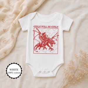 Puede incluir: Un body de beb&eacute; blanco de manga corta con un estampado gr&aacute;fico rojo de una persona montando un drag&oacute;n entre nubes. El texto GIRLS WILL BE GIRLS est&aacute; impreso en rojo sobre la ilustraci&oacute;n. Una etiqueta circular indica 100% algod&oacute;n.
