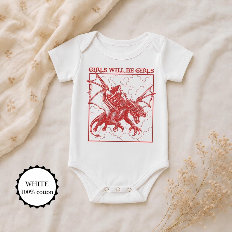 Puede incluir: Un body de beb&eacute; blanco de manga corta con un estampado gr&aacute;fico rojo de una persona montando un drag&oacute;n entre nubes. El texto GIRLS WILL BE GIRLS est&aacute; impreso en rojo sobre la ilustraci&oacute;n. Una etiqueta circular indica 100% algod&oacute;n.