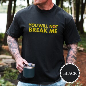 May include: Black crew neck t-shirt featuring the yellow text "YOU WILL NOT BREAK ME". A black circle with the word "BLACK" is in the lower right corner. A person is holding a dark blue mug. The shirt is a casual style.