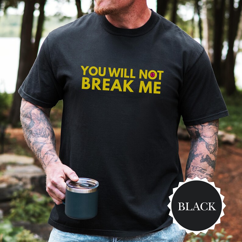May include: Black crew neck t-shirt featuring the yellow text "YOU WILL NOT BREAK ME". A black circle with the word "BLACK" is in the lower right corner. A person is holding a dark blue mug. The shirt is a casual style.