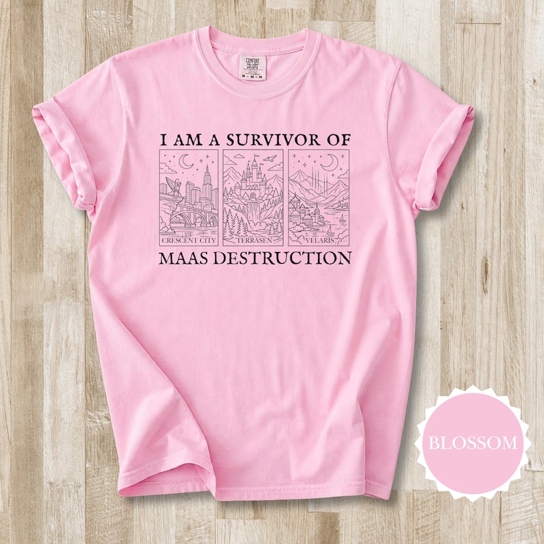 K&ouml;nnte beinhalten: Ein hellrosa T-Shirt mit hochgekrempelten &Auml;rmeln. Auf der Vorderseite befindet sich eine schwarze Grafik mit dem Text "I AM A SURVIVOR OF MAAS DESTRUCTION" und Illustrationen von Crescent City, Terrasen und Velaris.