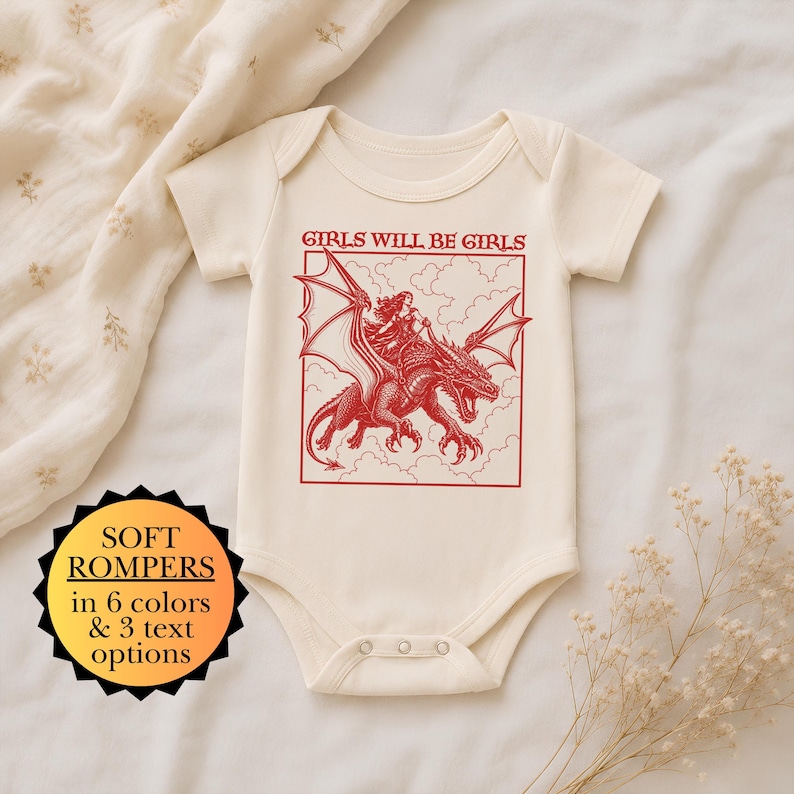 Puede incluir: Un body de manga corta color crema con un estampado gr&aacute;fico rojo de una persona montando un drag&oacute;n entre nubes. El texto GIRLS WILL BE GIRLS aparece sobre la ilustraci&oacute;n. Una insignia negra en forma de estrella muestra texto informativo.