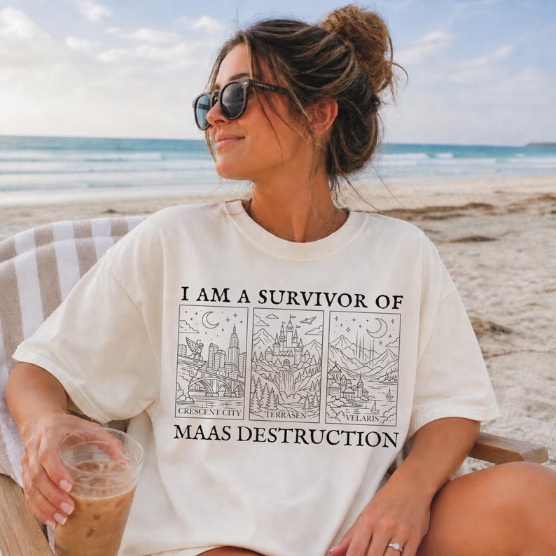 K&ouml;nnte beinhalten: Cremefarbenes T-Shirt mit dem Text "I AM A SURVIVOR OF MAAS DESTRUCTION" und drei illustrierten Tafeln. Die Tafeln zeigen Stadtlandschaften und Landschaften. Die Person auf dem Bild tr&auml;gt eine Sonnenbrille und sitzt am Strand.