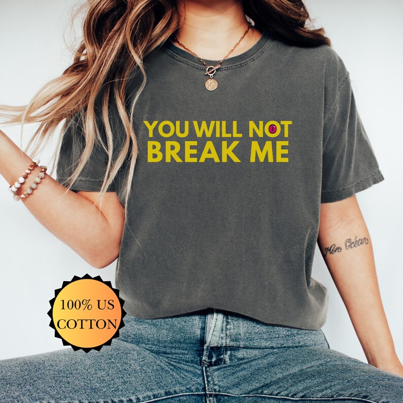 May include: Dark grey t-shirt with the text "YOU WILL NOT BREAK ME" in yellow. The shirt also has a small red dot. The shirt is made of 100% US cotton.